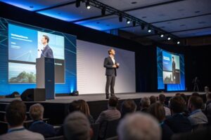 A confident speaker delivering a presentation on stage to an attentive audience.